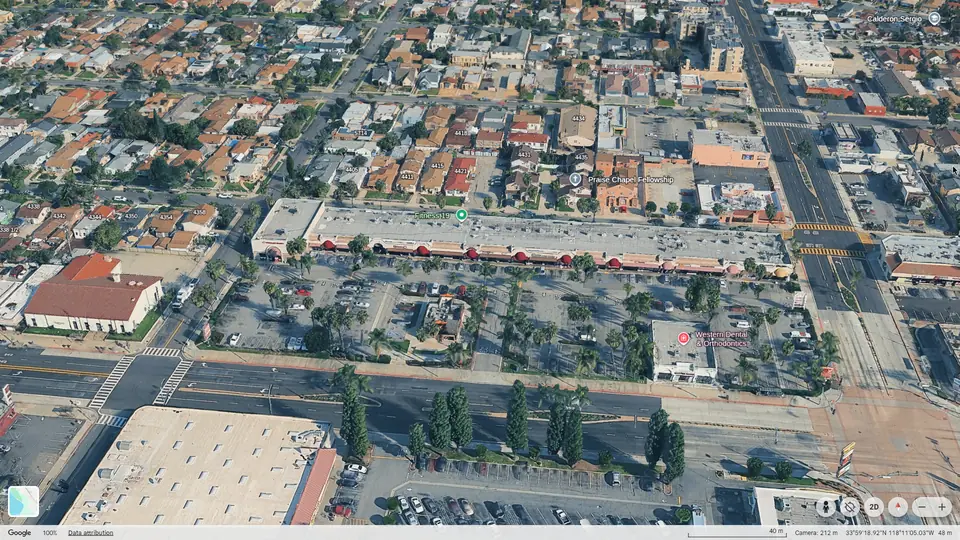 Google Earth 3D view of Maywood Village Center from E Slauson Ave, showing the single-story strip of storefronts with red awnings behind a palm-lined parking lot